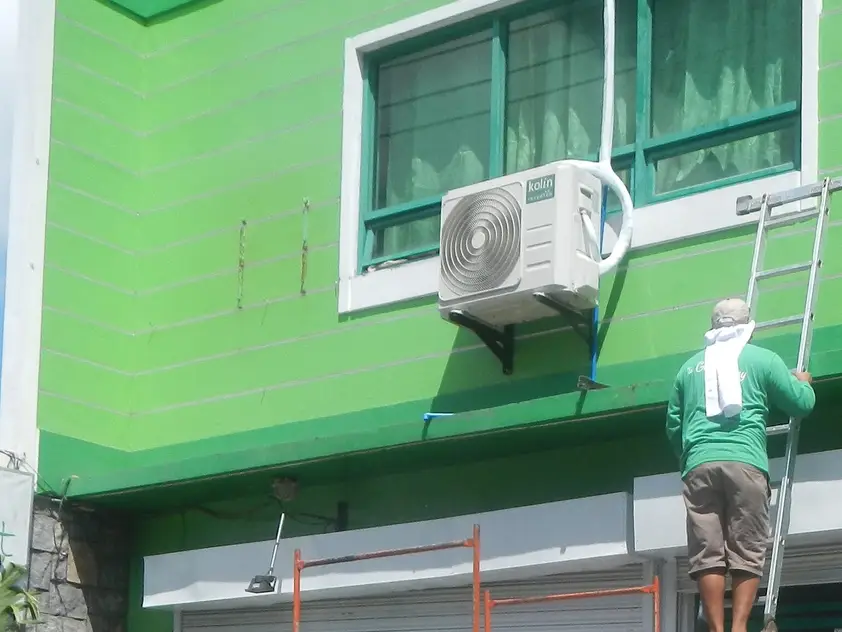 Instalador subido a una escalera junto a la unidad exterior de un aire acondicionado, ya fijada a la fachada con un soporte metálico
